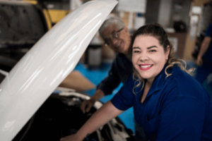 Raising the Bar: Your Guide to Creating a Great Shop Culture by Chris Cotton, AutoFix Auto Shop Coaching. Image of a smiling woman with a slightly old man in the background, working under the hood of a car.
