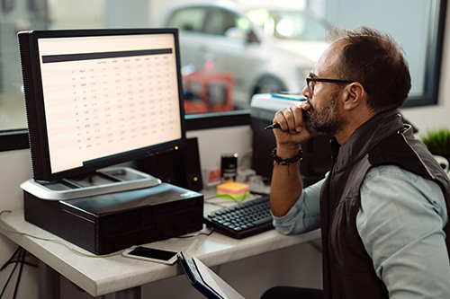 Importance of Precise Income Statements for Tax Season and Beyond | AutoFix Auto Shop Coaching. Image of an auto shop’s foreman using desktop PC in the office and making financial reports.