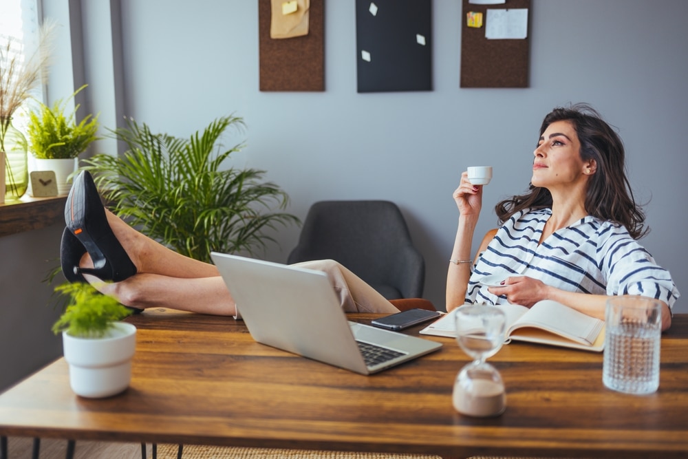 Mid-Year Reflection for Auto Repair Shops by AutoFix-Auto Shop Coaching. A woman relaxing at her desk with her feet up, holding a cup of coffee and reflecting, symbolizing the importance of taking a break to assess the performance and future goals of an auto repair shop for a successful second half of the year.