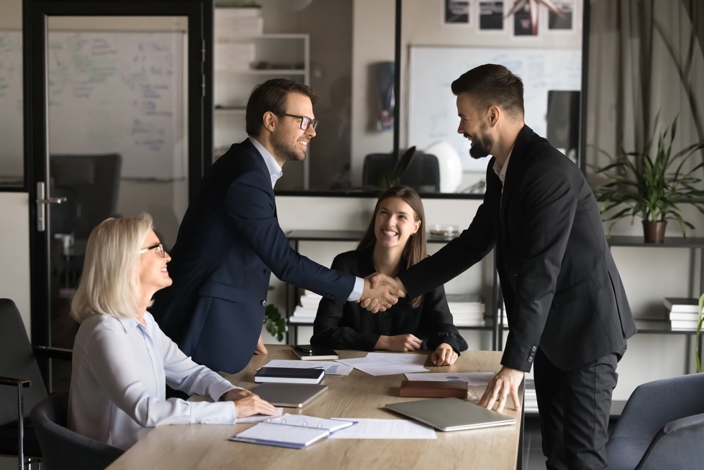 Commitment business coach by AutoFix- Auto Shop Coaching. A business meeting with two men in suits shaking hands, symbolizing commitment and partnership, while two women observe and smile, representing a successful, engaged collaboration.