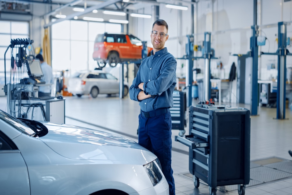  Unlocking financial success with AutoFix Auto Shop Coaching. Financially successful auto mechanic posing in a car service. Wearing jeans, shirt, and glasses with arms crossed.