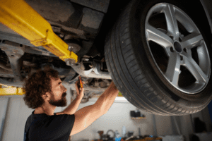 Auto technician, auto shop coaching by AutoFix Auto Shop Coaching. Auto technician performing undercar inspection on a lifted vehicle, checking suspension and tire components with a flashlight during routine auto repair service in a professional repair shop.