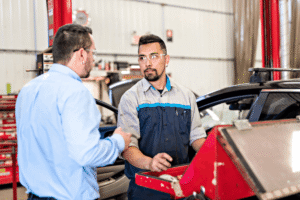 Auto repair technician, auto shop coaching at AutoFix Auto Shop Coaching. Image of a technician speaking with an owner beside a vehicle and tool chest inside a service shop, supporting clear repair communication.